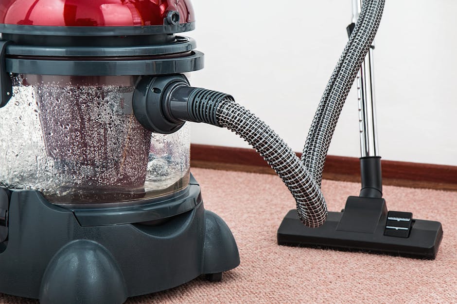 A professional cleaner dressed in a full protective suit, including a white coverall, face mask, gloves, and shoe covers, is performing deep carpet cleaning in a bright residential living room. The cl